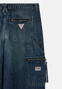 Dunkelblaue Denim-Cargohosen mit mehreren Taschen, einschließlich einer Reißverschlusstasche. Mit Markenlogos und kontrastierenden roten und weißen Akzenten.