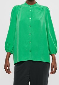 Groene blouse met lange mouwen en knopen aan de voorkant, gedragen door iemand in een zwarte broek, met een ontspannen pasvorm en gathered mouwen.