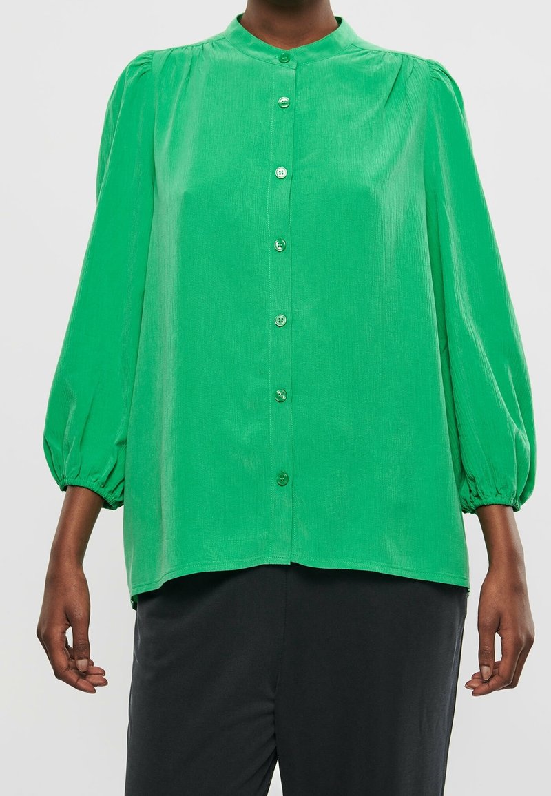 Groene blouse met lange mouwen en knopen aan de voorkant, gedragen door iemand in een zwarte broek, met een ontspannen pasvorm en gathered mouwen.