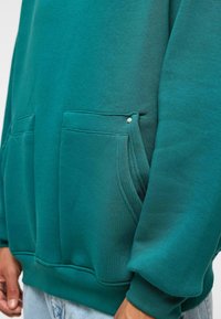 Teal hoodie met twee voorzakken, zachte stof, ribgebreide cuffs en zoom. Heeft een klein drukknoopdetail op de rand van de zak.