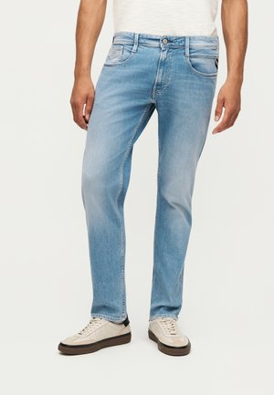 Mann trägt hellblaue Jeans mit geradem Schnitt und weiße Sneakers, steht vor einem schlichten Hintergrund mit entspannt an den Seiten hängenden Händen.