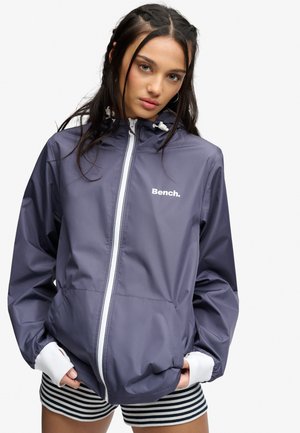 Ung kvinde iført en navy Bench-jakke med hvid lynlås og stribede shorts, stående mod en ensfarvet hvid baggrund.