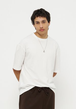 Mladý muž s kučeravými vlasmi nosiaci jednoduché biele voľné tričko, tmavo hnedé nohavice so sťahovacou šnúrkou a strieborný prívesok na bielom pozadí.