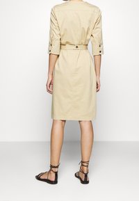 Khakifarbene Hemdblusenkleid mit Dreiviertelärmeln, tailliertem Schnitt und Knopfakzenten. Kombiniert mit schwarzen flachen Sandalen, die ein minimalistisches Design aufweisen.