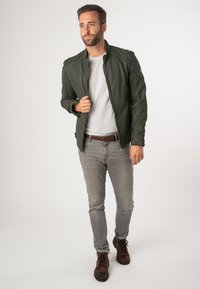 Olijfgroene leren jas met ritssluiting, gestructureerd oppervlak en schouderdetails, gedragen over een lichtgrijs t-shirt en grijze spijkerbroek.