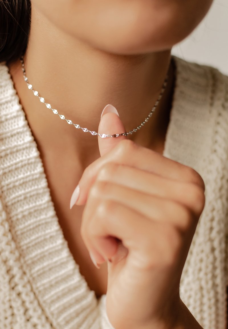 Silberne herzförmige Choker mit einem filigranen Kettendesign, das auf der Haut liegt. Die Hand des Models berührt sanft die Halskette.