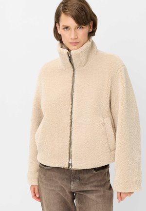 Woman wearing a beige fleece jacket with a high collar and front zipper, paired with brown pants, against a plain background.