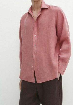 Mand iført en løs, langærmet, knaplukket pink hørskjorte med mørke bukser, stående mod en ensfarvet lys baggrund.