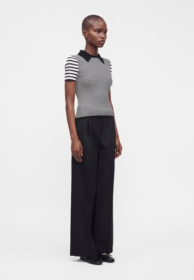 Personne debout portant un pantalon noir à jambes larges et un haut à manches courtes rayé noir et blanc avec un col noir sur un fond uni.