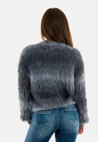 Chaqueta de piel sintética gris con un corte holgado, superficie texturizada y mangas largas, combinada con jeans azules ajustados que cuentan con bolsillos traseros distintivos.