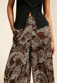 Pantalons larges à motifs marron avec un design paisley blanc, présentant une taille haute et un tissu lisse et léger. Chemise noire avec des boutons.