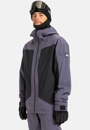 Homme portant une veste de ski noire et grise, un pantalon assorti, des gants noirs, un bonnet noir et des lunettes de ski sur le front, sur un fond uni.
