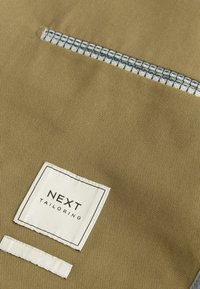 Tan stof med en glat tekstur, prydet med en hvid etikette med "NEXT TAILORING" og en dekorativ lynlås i blå og hvid.