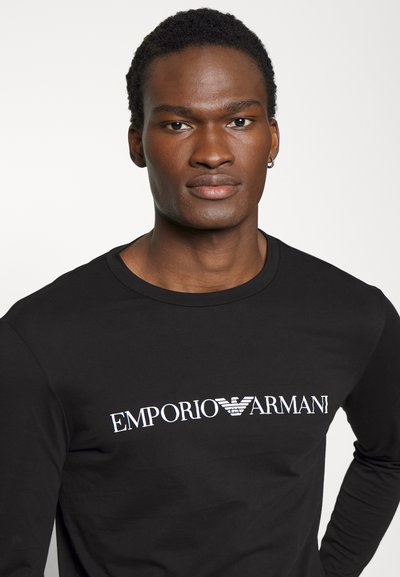 Crna dug sleeves košulja od mekog materijala, s bijelim "Emporio Armani" logotipom preko prsa. Jednostavan dizajn, okrugli izrez.