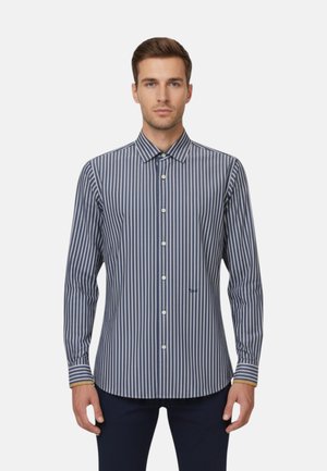 Uomo che indossa una camicia a maniche lunghe con bottoni e righe verticali blu e bianche, in piedi davanti a uno sfondo chiaro e uniforme.