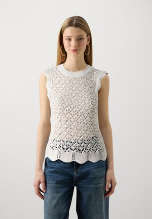 Jersey sin mangas blanco de punto con un patrón de encaje, dobladillo con forma de concha y cuello de canalé, llevado sobre unos jeans azules.