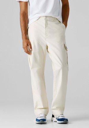 Mann trägt cremefarbene Cargo-Hose, weißes T-Shirt und weiße Sneaker mit blauen Akzenten, steht vor einem schlichten grauen Hintergrund.