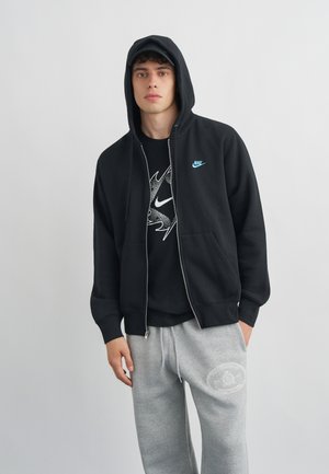Młody mężczyzna w czarnej bluzie Nike z kapturem i zamkiem, czarnej koszulce z grafiką oraz jasnoszarych spodniach dresowych z białym emblematem.