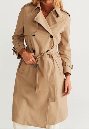 Nainen, joka pukeutunut beigeen kaksiriviseen trenssitakkiin, jossa on mustat napit, solmittava vyö ja soljilla varustetut hihansuut, yllä ruskea pooloneule.