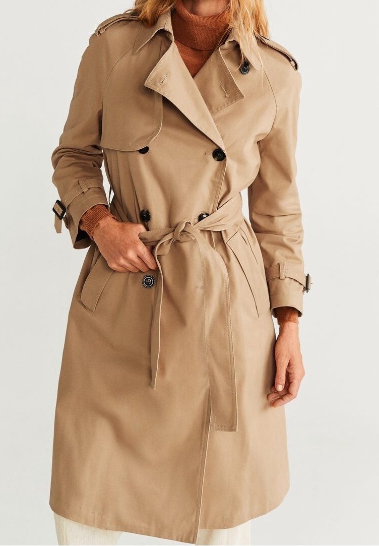 Femme portant un trench-coat beige à double boutonnage avec des boutons noirs, une ceinture nouée et des manches à boucles, par-dessus un pull à col roulé marron.