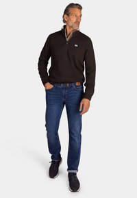 Donkerbruine trui met rits, blauwe spijkerbroek en donkere sneakers. De outfit heeft een casual pasvorm met een gestructureerde brei en een klein logo op de borst.