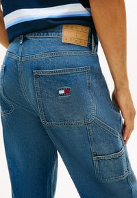 Man draagt blauwe Tommy Jeans spijkerbroek met meerdere zakken en merkpatches op de achterzak en tailleband.