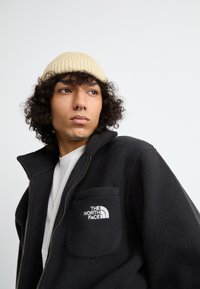 Must mustan fleece-jakk, millel on lukuga sulgumine, ees tasku ja The North Face logo. Seljas valge särk, koos beeži ribaga mütsiga.