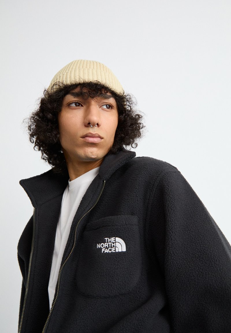 Veste en polaire noire avec fermeture éclair, dotée d'une poche ceinturante et du logo The North Face. Portée sur une chemise blanche, accompagnée d'un bonnet beige à côtes.
