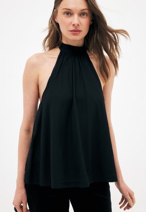 Kobieta w czarnej, bezrękawnikowej bluzce z odkrytymi ramionami i luźnym fasonie, stojąca na białym tle, z lekko rozwianymi włosami na bok.