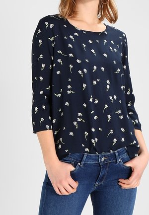 Kvinde iført en mørkeblå bluse med små hvide blomsterprint og trekvartede ærmer, kombineret med blå jeans, hænderne i lommerne.