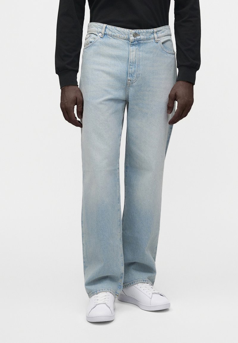 Personne portant un jean bleu clair à jambes larges, des baskets blanches et un t-shirt noir à manches longues sur un fond uni.