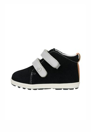Zapato negro para niño pequeño con tiras dobles de velcro blancas, costuras blancas y suela blanca con banda de rodadura negra.