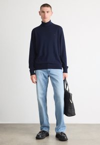 Marinefarbener Rollkragenpullover aus weichem Strick, kombiniert mit hellblauen gerade geschnittenen Jeans. Schwarze Tasche in einer Hand, klobige schwarze Schuhe. Minimalistischer Stil.