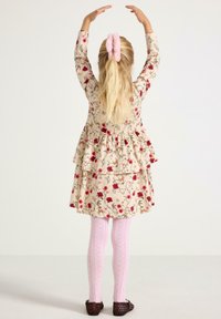 Młoda dziewczynka z długimi blond włosami związanymi różową gumką, ubrana w kwiecistą sukienkę, różowe rajstopy i ciemne buty, z rękami uniesionymi nad głową.