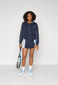 Marinblå atletisk zip-jacka med vita detaljer, tillsammans med en mörk klänning med ljusblå sidopaneler. Håller en tennisracket och en boll.
