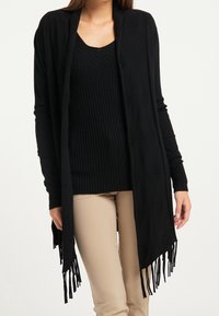 Long cardigan noir avec texture côtelée, ouvert à l'avant, et ourlet à franges. Porté sur un top noir côtelé et un pantalon beige clair.