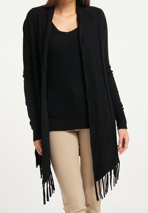 Long black cardigan with ribbed texture, open front, and fringed hem. Worn over a black ribbed top and light beige pants.