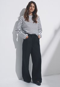 Camisa de manga larga blanca con rayas azul marino combinada con pantalones anchos de color azul marino oscuro. El modelo está de pie con las manos en los bolsillos.