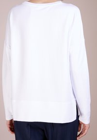 Osoba v jednoduché bílé košili s dlouhými rukávy, viděná zezadu, v kombinaci s tmavě modrými kalhotami, stojící na neutrálním pozadí.