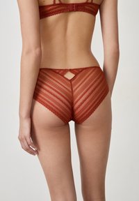 Slip rosso in pizzo con un motivo a righe, caratterizzato da un piccolo dettaglio a forma di chiave sul retro e bordi ondulati lungo la vita e le aperture delle gambe.