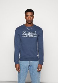 Mornarsko plavi dukserica s rebrastim manžetama i rubom, koja na prsima ima bijeli natpis "Original Claim Your Denim Rights Blend".