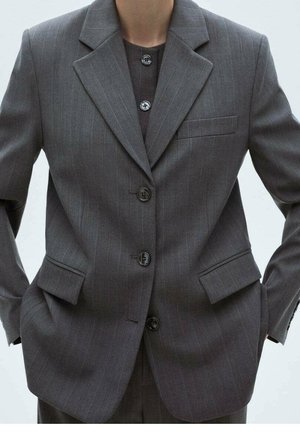 Personne portant une veste de costume à fines rayures gris foncé avec poches à rabat et trois boutons, mains dans les poches, face à l'avant.