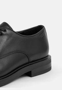 Chaussure en cuir noir avec un bout rond, texture lisse, couture discrète et un talon bas empilé. Marque au niveau du talon.