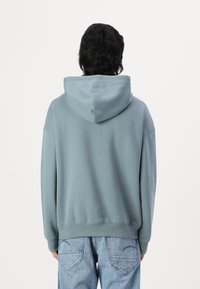 Felpa con cappuccio di colore azzurro chiaro, con vestibilità comoda, texture morbida, spalle scese e polsini a coste; abbinata a jeans in denim scolorito.