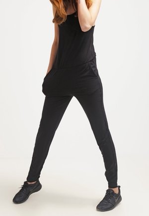 Kobieta ubrana w czarny bezrękawnik i czarne trampki, stojąca z nogami rozstawionymi na boki, jedną ręką w kieszeni.