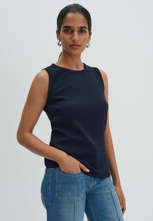 Donna con capelli scuri, indossa una canotta blu scuro e jeans blu, in piedi con una mano in tasca contro uno sfondo grigio uniforme.