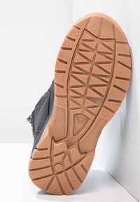 Suola di un boot scuro con un battistrada in gomma marrone spesso e con motivo, appoggiata su una superficie bianca.