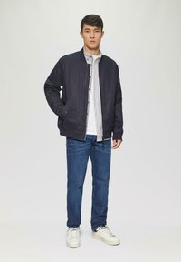 Chaqueta de piloto en color marino con puños acanalados, una camisa blanca con rayas debajo, jeans de color azul oscuro y zapatillas blancas. Diseño simple y casual.