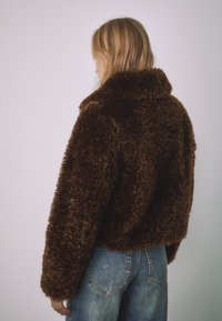 Hnedá huňatá bunda s vysokým golierom a spustenými ramenami, kombinovaná so svetlomodrými rifľami. Má jemnú štruktúru a skrátený strih.