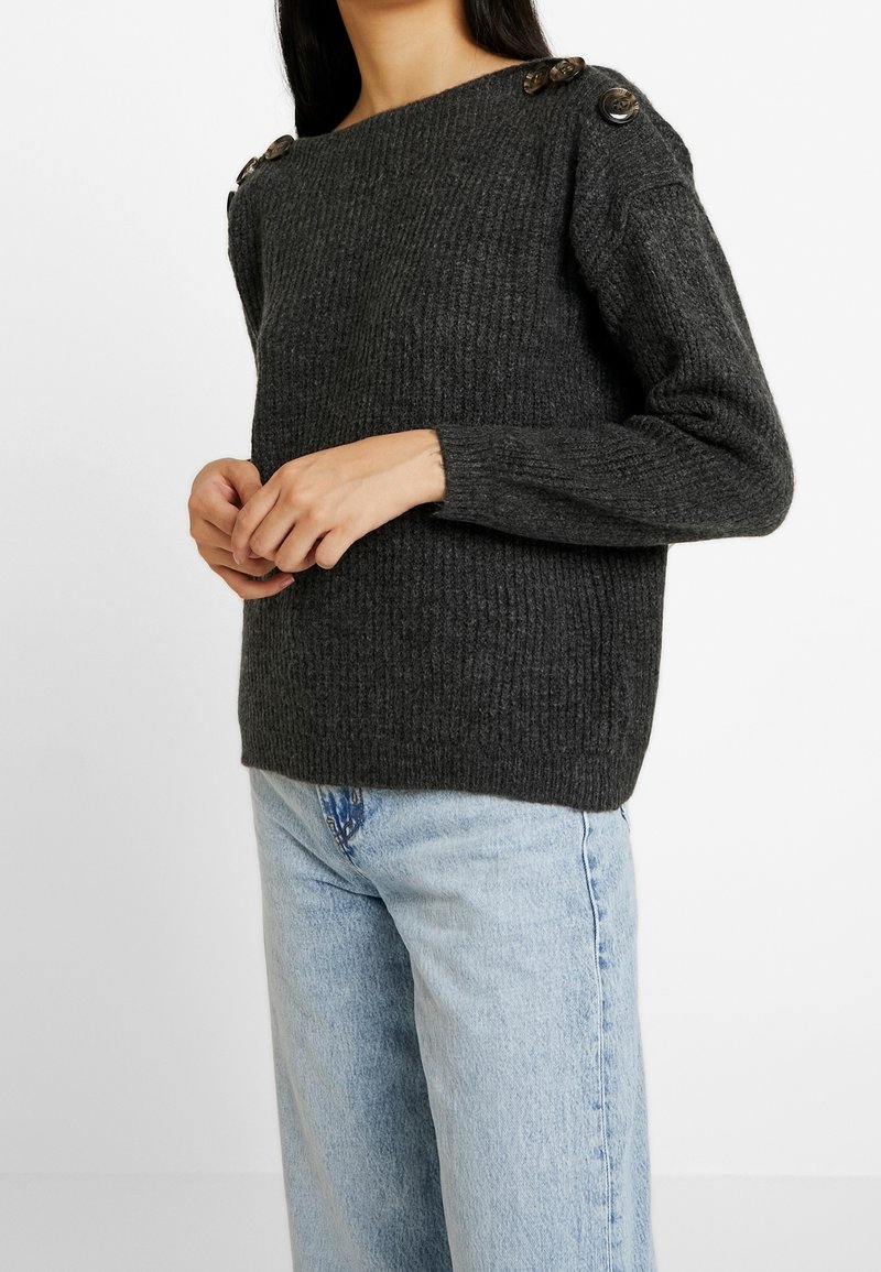 Femme portant un pull en maille côtelée gris foncé avec de grands boutons sur les épaules et un jean en denim bleu clair, debout devant un fond blanc.